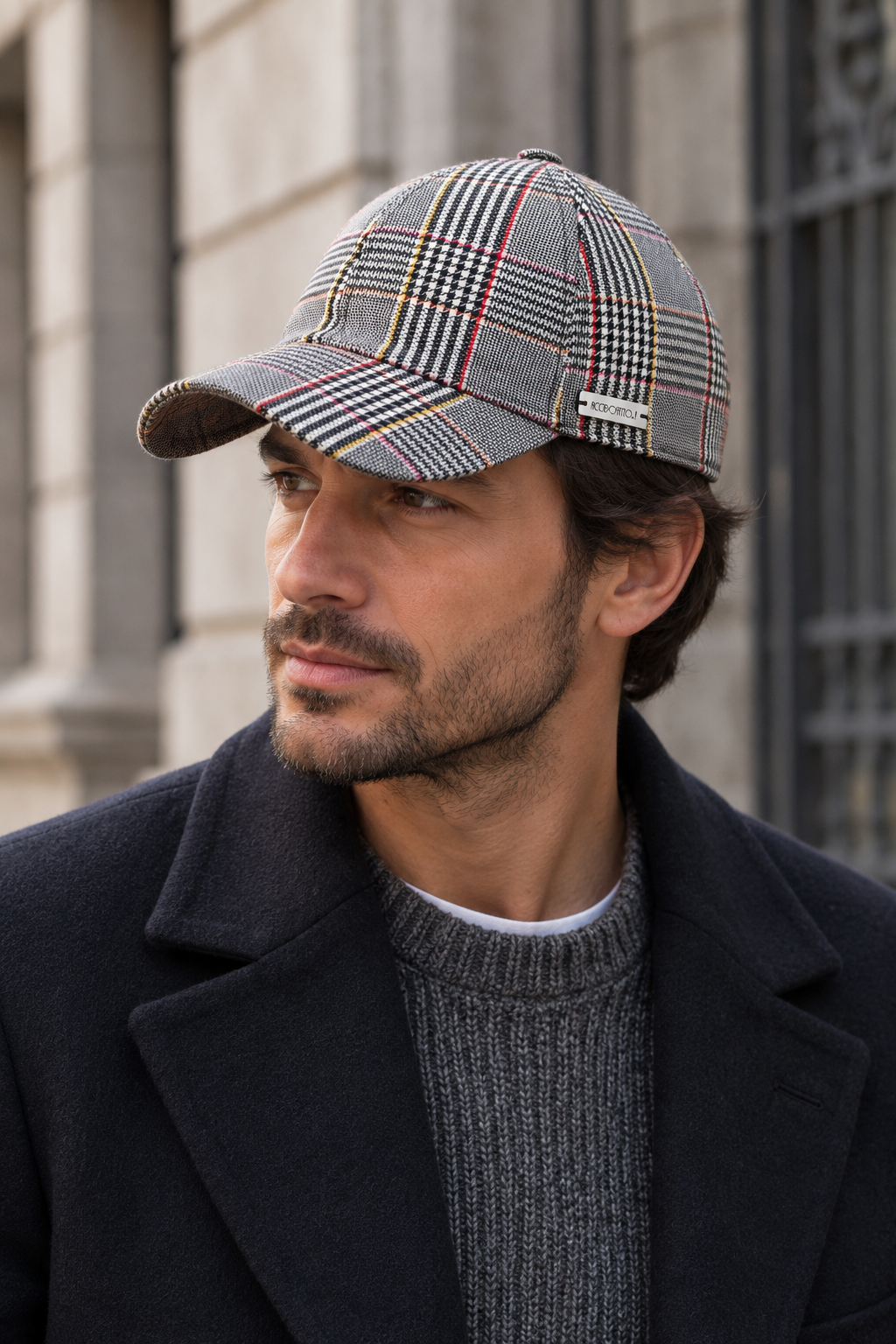Cappello baseball Bry del brand Accapofitto, in tessuto fantasia Principe di Galles, con alcune righe sul rosso e sul giallo, indossato da un modello con cappotto blu e maglione grigio