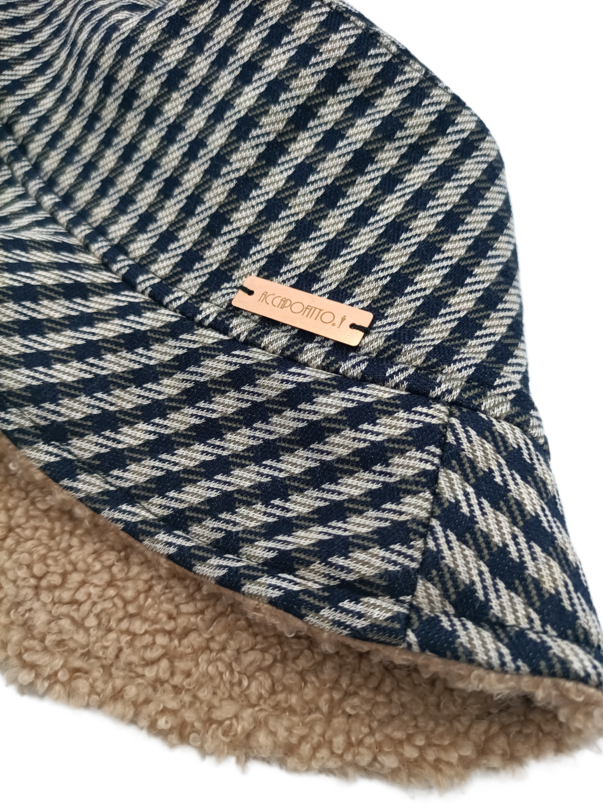 Cappello bucket hat Mabel del brand Accapofitto, realizzato con tessuto a quadretti blu e beige all'esterno e con tessuto teddy color cammello all'interno. Dettaglio etichetta logo color rame