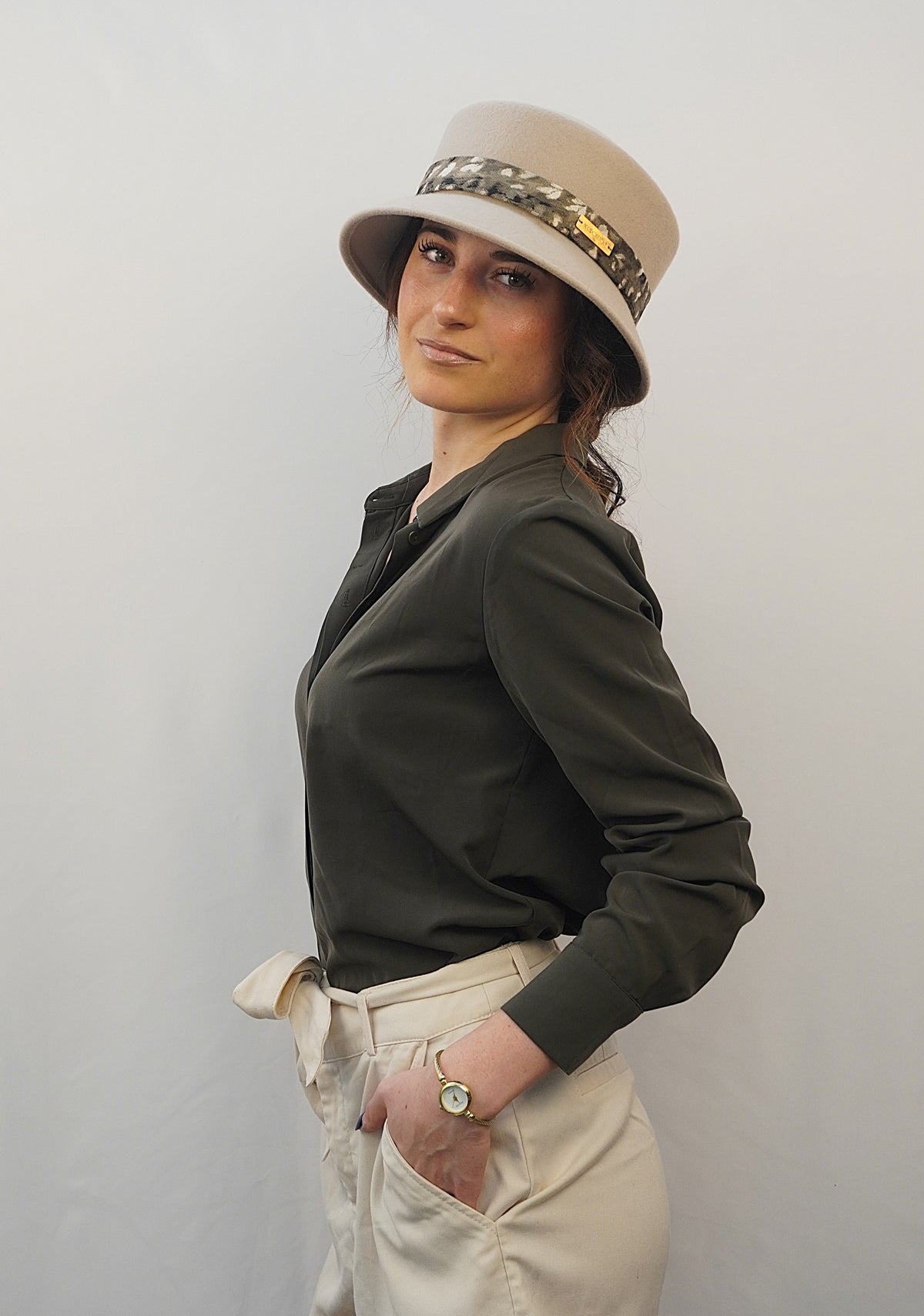 Cappello Edith del brand Accapofitto: bucket hat di feltro beige con capino sbalzato e decorato con fascia a fantasia sui toni del verde militare del nero e del bianco. Indossato da una modella con camicia verde epantaloni beige in uno studio fotografico