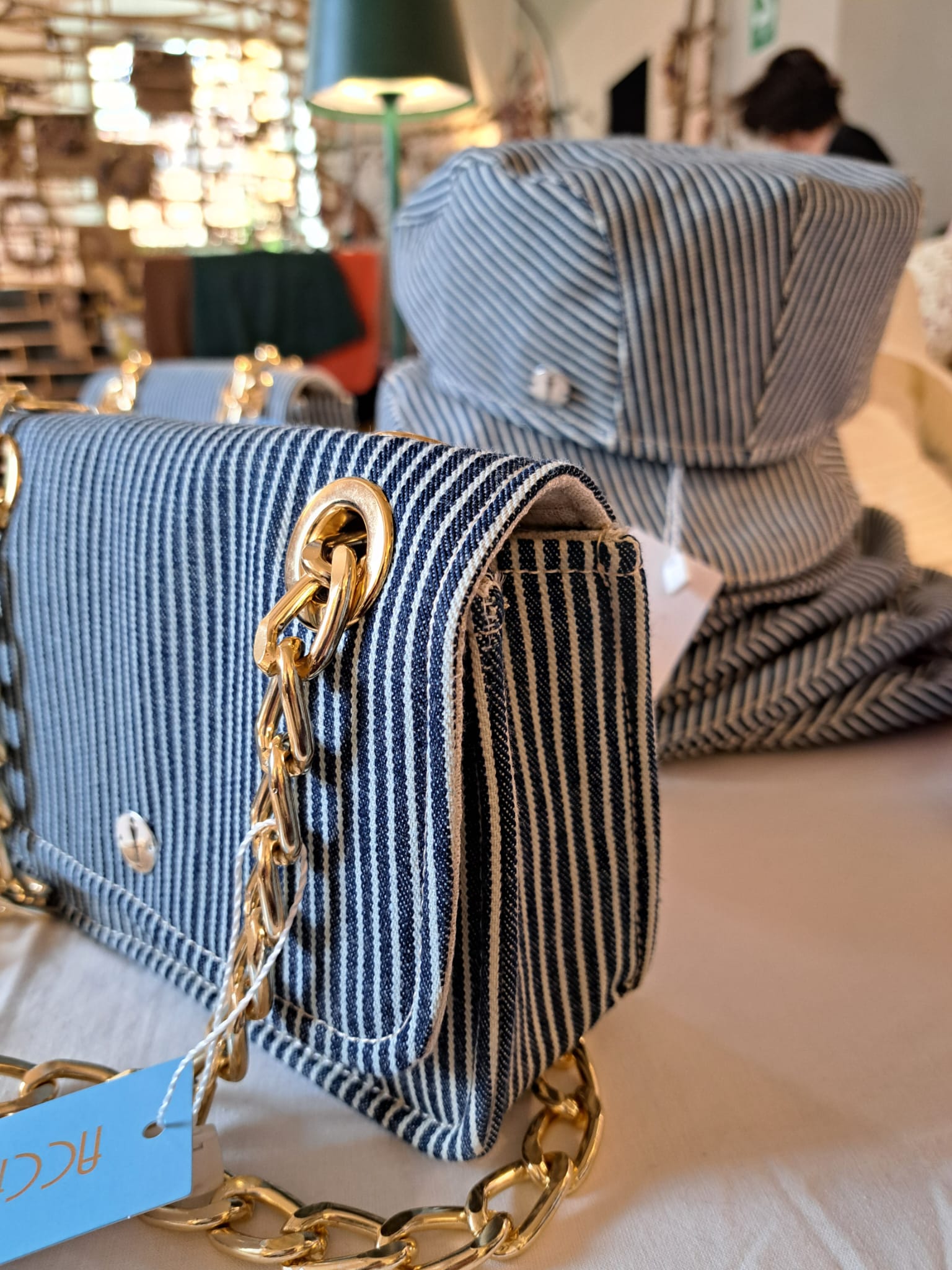 Foto espositiva della pochette Denny del brand Accapofitto, in denim rigato bianco con catena dorata, e sullo sfondo la coppola abbinata